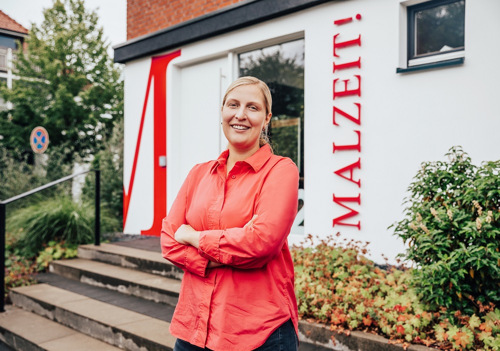 Eine lächelnde Frau in einem roten Hemd steht mit verschränkten Armen vor einem Gebäude, an dessen Wand in großen roten Buchstaben MALZEIT! steht. Im Hintergrund sind Treppen, Pflanzen und Bäume zu sehen.