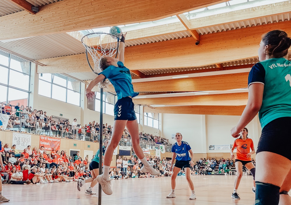 Eine Netzballspielerin in Blau springt hoch, um den Ball während eines Spiels in einer überfüllten Sporthalle ins Netz zu schießen, während Zuschauer und andere Spieler das Geschehen beobachten.