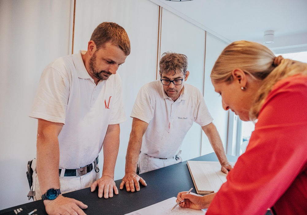 Als Frau im Handwerk lehnt sich eine Frau in rotem Hemd über den Tisch, um Dokumente zu unterschreiben, während zwei Männer in weißen Arbeitsuniformen aufmerksam zusehen. Die Szene spielt in einem Büro oder Besprechungsraum.
