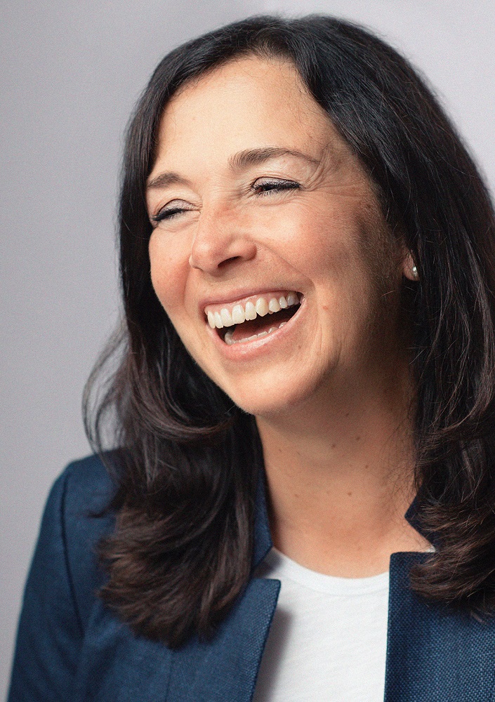 Sabine Logemann strahlt Selbstentfaltung aus: Eine Frau mit langem dunklem Haar, im blauen Blazer und weißen Top, lacht und lächelt, während sie leicht zur Seite blickt - vor schlichtem, hellem Hintergrund.