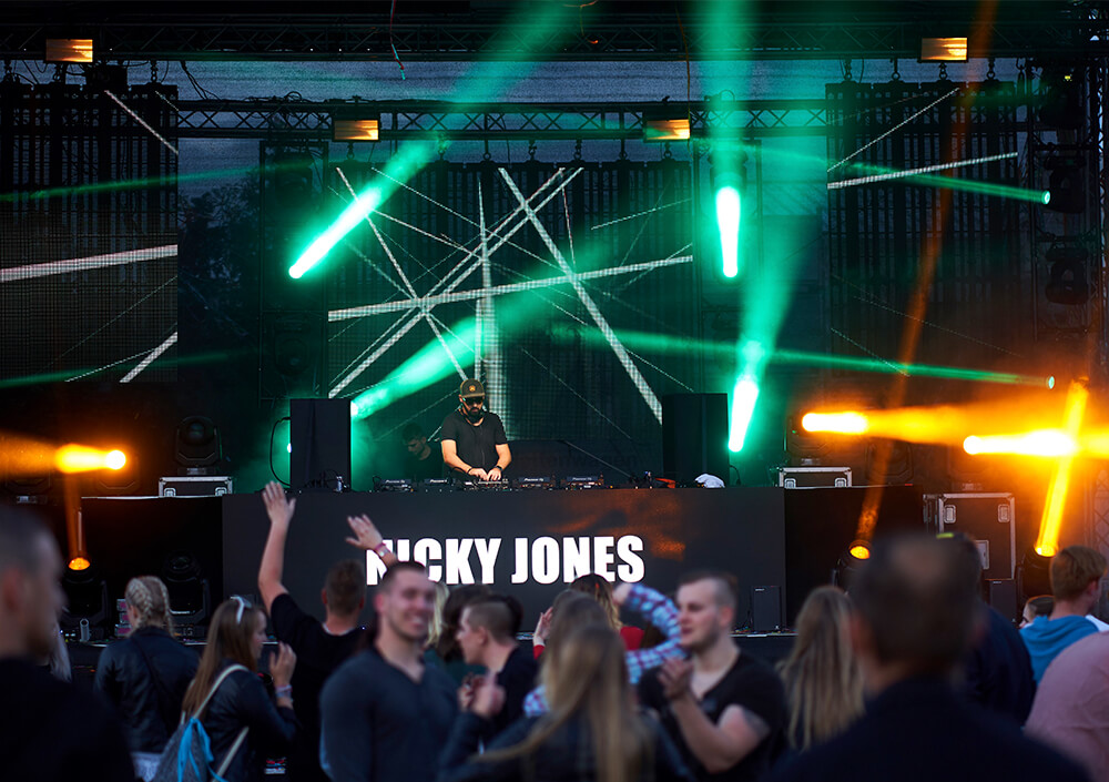 Ein DJ im Club legt auf der Bühne mit leuchtend grünen und orangefarbenen Lichtern und Lasereffekten auf, während die Menge tanzt und die Hände hebt. Auf dem DJ-Pult steht in großen weißen Buchstaben der Name NICKY JONES.