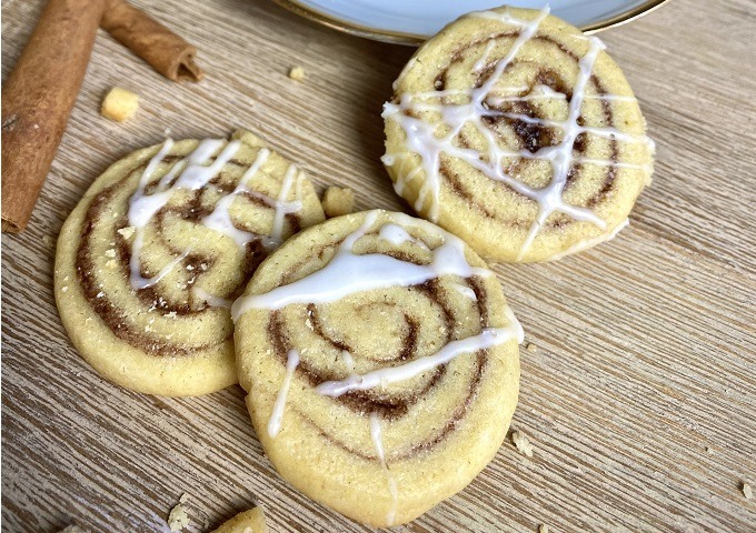 Drei Zimtplätzchen, die mit weißem Zuckerguss beträufelt sind, werden auf einer Holzfläche präsentiert, die an klassische Vanille-Plätzchen erinnert. Im Hintergrund sind Zimtstangen und ein Teil eines weißen Tellers zu sehen.