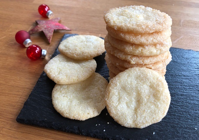 Ein Stapel goldener Vanille-Plätzchen und mehrere lose Plätzchen sind auf einer schwarzen Schiefertafel angeordnet. Festliche rote Ornamente und ein roter Stern sorgen für weihnachtliche Stimmung auf der Holzfläche neben den Plätzchen.