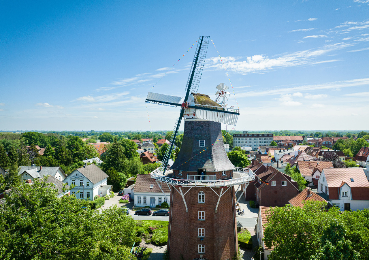 Eine traditionelle Windmühle mit Backsteinsockel steht inmitten von Häusern und Grünanlagen unter einem strahlend blauen Himmel in der malerischen Stadt Varel und ist ein echter Lieblingsort für Besucher und Einheimische gleichermaßen.