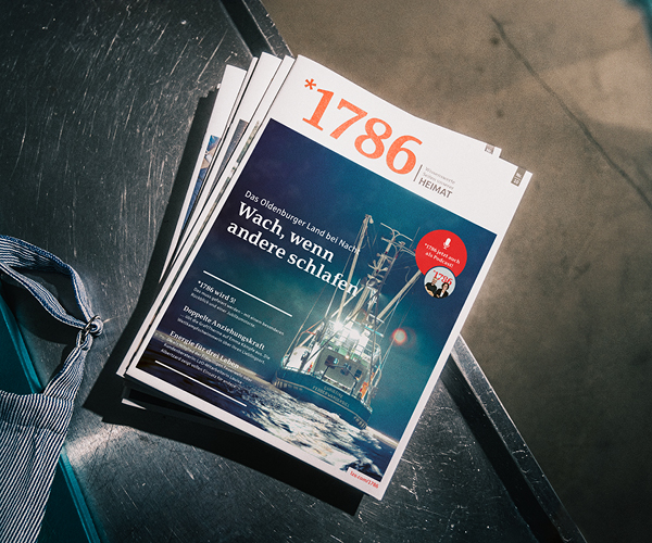 Ein Stapel Zeitschriften mit dem Titel 1786, ein Online Magazin, liegt auf einer Metallfläche. Das Cover zeigt ein Schiff auf nächtlicher See mit der Schlagzeile "Wach, wenn andere schlafen". Am Rand ist ein Teil einer blau gestreiften Tasche zu sehen.