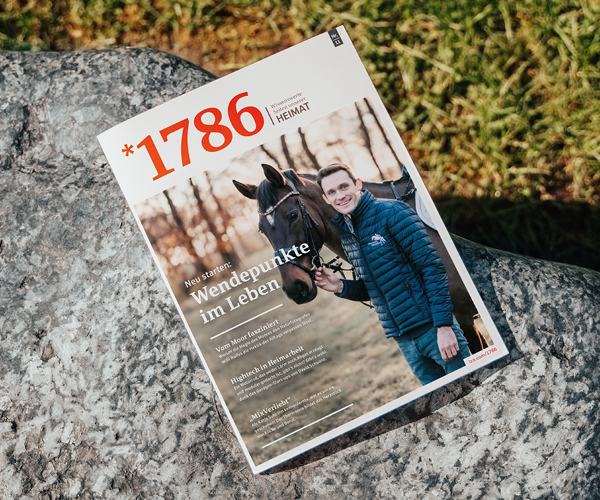 Eine Ausgabe des Online Magazins *1786 liegt auf einer steinernen Oberfläche. Sie zeigt einen lächelnden Mann in einer blauen Jacke, der die Zügel eines braunen Pferdes hält. Im Hintergrund sind Gras und Sonnenlicht zu sehen.