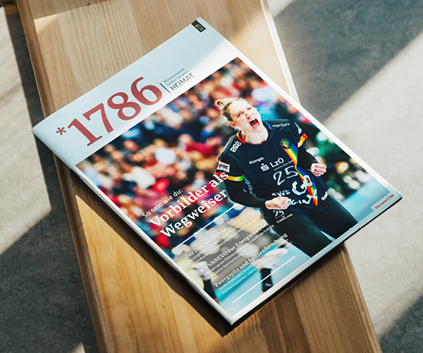 Ein Online Magazin *1786 mit dem Titel "1786" ruht auf einer Holzbank. Das Titelbild zeigt eine jubelnde Sportlerin in einem schwarzen Trikot, hinter ihr eine verschwommene Menge und mehrere deutsche Schlagzeilen.