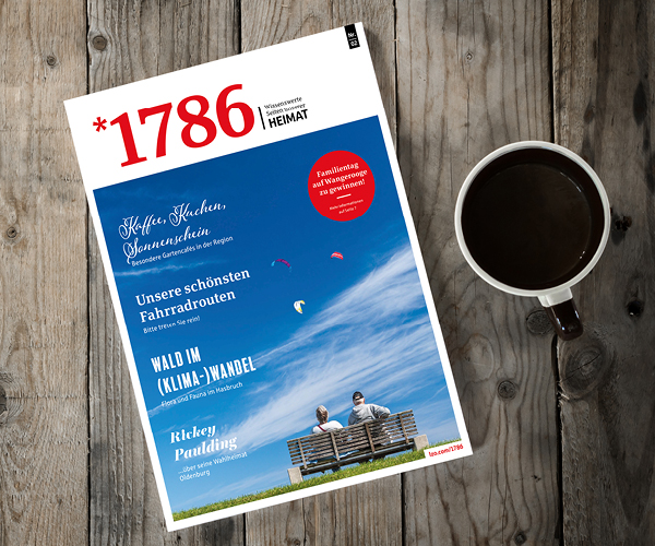 Ein Online Magazin *1786 liegt auf einem Holztisch neben einer Tasse schwarzen Kaffees. Das Titelbild zeigt einen blauen Himmel mit Wolken, Menschen auf einer Bank und bunte Flugdrachen. Die Überschrift hebt lokale Besonderheiten und Fahrradrouten hervor.