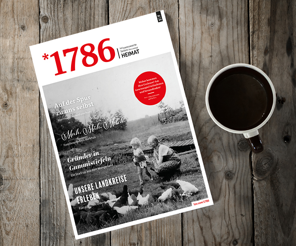 Eine Ausgabe des Online Magazins *1786 mit einem Schwarz-Weiß-Foto vom Bauernhof auf dem Cover liegt auf einem Holztisch neben einer Tasse schwarzen Kaffees. Das Magazin enthält deutschen Text und einen roten Kreis, der ausgewählte Inhalte hervorhebt.