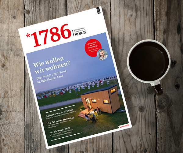 Eine Ausgabe des Online-Magazins *1786 liegt auf einem Holztisch neben einer Tasse Kaffee. Das Titelbild zeigt ein modernes kleines Haus, Strandkörbe am Meer und einen deutschen Text über zukünftige Wohntrends.
