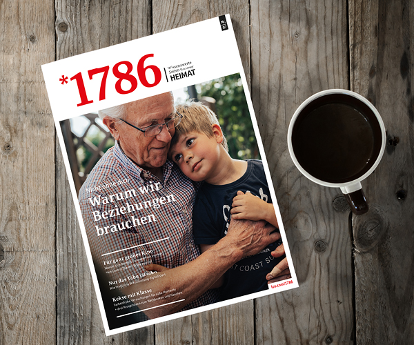 Eine Tasse Kaffee steht neben einem Online Magazin *1786, dessen Titelbild einen älteren Mann zeigt, der einen kleinen Jungen umarmt. Auf dem Holztisch prangt die Schlagzeile "1786" und ein deutscher Text über Beziehungen.