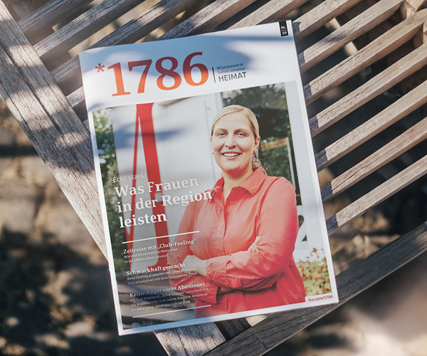 Eine Zeitschrift mit dem Titel Online Magazin *1786 Heimat liegt auf einer Holzbank. Das Titelbild zeigt eine lächelnde Frau in einem roten Hemd, die im Freien steht, mit einem deutschen Text über den Beitrag der Frauen in der Region.