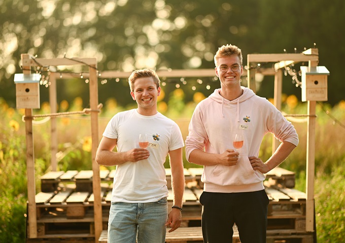 Zwei junge Männer stehen im Freien auf einem Feld, lächeln und halten Gläser mit Roséwein in der Hand. Sie tragen legere Kleidung und stehen vor einer mit Lichterketten geschmückten Holzkonstruktion mit Sonnenblumen und Grünpflanzen im Hintergrund.