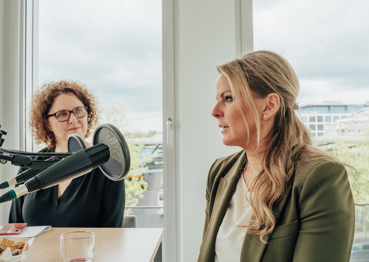 Zwei Frauen sitzen an einem Tisch mit Mikrofonen und nehmen einen Podcast oder ein Interview für *1786 trifft auf: Eva Högl. Eine Frau mit blondem Haar spricht, während die andere, mit lockigem Haar und Brille, aufmerksam zuhört, während die Lichter der Stadt durch große Fenster scheinen.