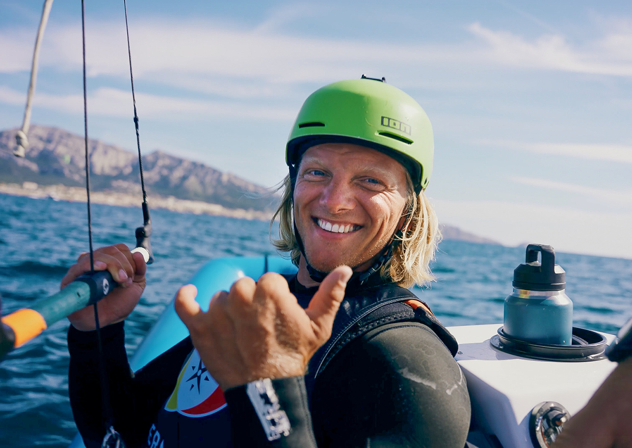 Eine lächelnde Person mit langen blonden Haaren, die einen grünen Helm und einen Neoprenanzug trägt, gibt ein Shaka-Zeichen, während sie auf dem Meer segelt, mit Bergen und einem blauen Himmel im Hintergrund -*1786 trifft: Eva Högl schwingt sich auf die Wellen.