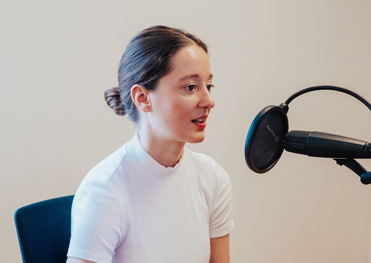 Eine junge Frau mit zurückgebundenen braunen Haaren und weißem Oberteil spricht in ein Mikrofon mit Popfilter in einer Tonstudioumgebung für 1786 trifft: Eva Högl.