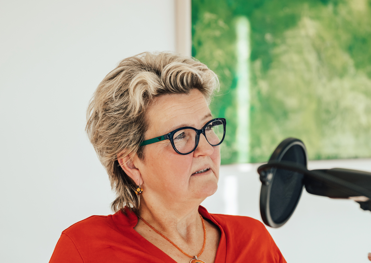 Eine Frau mit kurzen, gewellten grauen Haaren und Brille, die ein rotes Oberteil trägt, sitzt vor einem Mikrofon. Sie scheint zu sprechen oder zuzuhören, mit einem grünen Gemälde im Hintergrund - Teil der Serie *1786 trifft: Eva Högl-Serie.