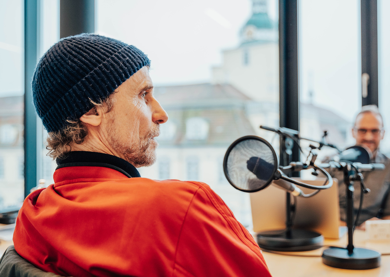 Ein Mann mit roter Jacke und marineblauer Strickmütze sitzt an einem Tisch mit einem Mikrofon und spricht während der Podcast-Aufnahme "1786 trifft: Eva Högl". Im Hintergrund ist eine weitere Person mit Brille zu sehen. Große Fenster zeigen Gebäude im Freien.