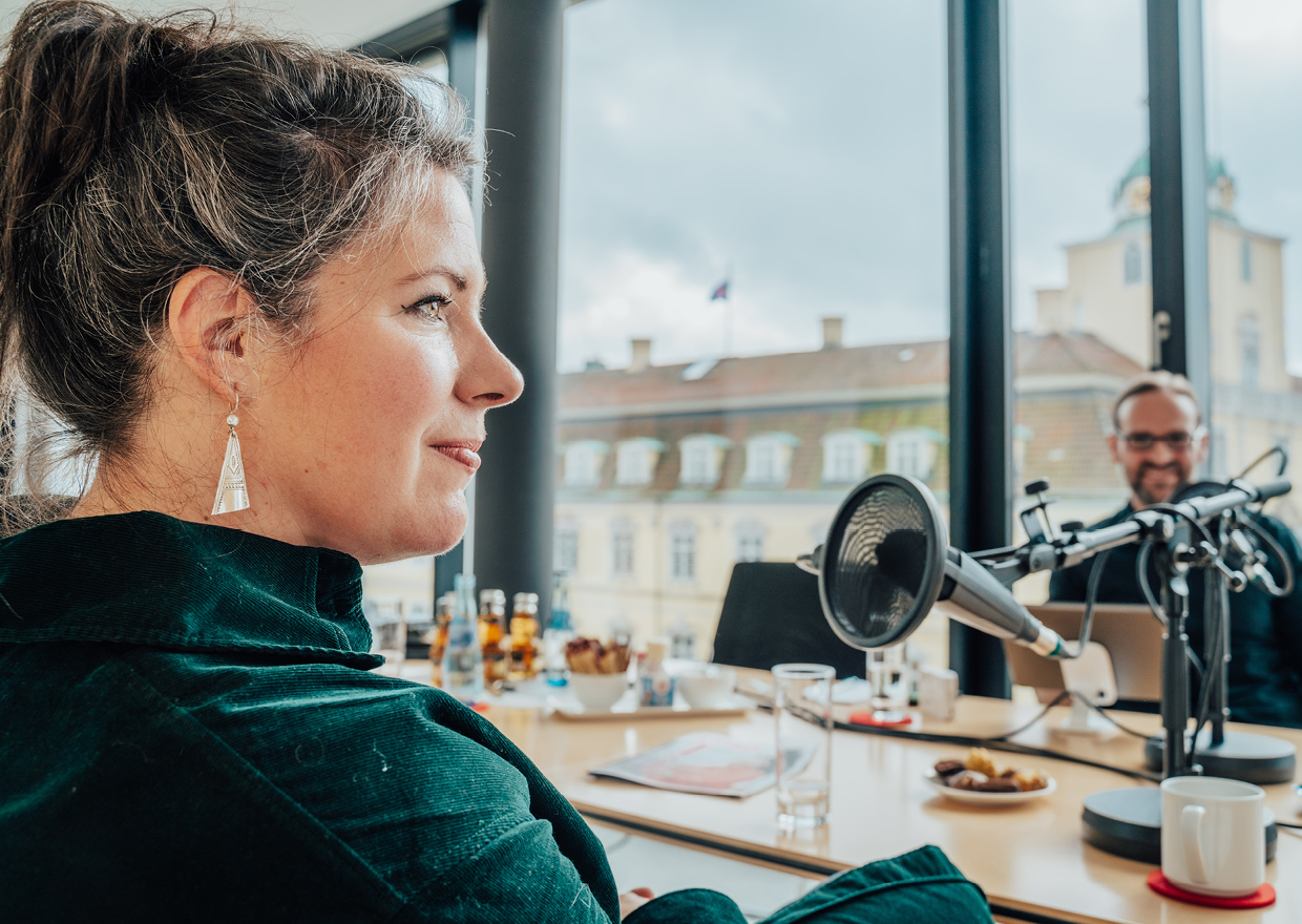 Eine Frau mit graumeliertem Haar sitzt in einem hellen, modernen Raum vor einem Mikrofon und bereitet sich auf 1786 trifft vor: Eva Högl. Ihr gegenüber sitzt ein Mann mit einem Laptop und lächelt. Auf dem Tisch stehen Getränke und Snacks.