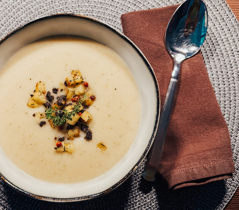 Eine Schüssel mit einer cremigen Suppe mit gewürfeltem Gemüse und Kräutern, die auf einem gewebten Tischset neben einer braunen Serviette und einem glänzenden Löffel steht.