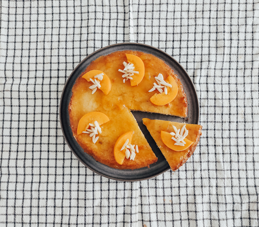 Auf einem dunklen Teller liegt ein runder Kuchen mit Pfirsichscheiben und weißen blumenförmigen Verzierungen. Eine Scheibe ist angeschnitten und leicht abgetrennt. Der Teller steht auf einem weißen Tuch mit einem schwarzen Gittermuster.