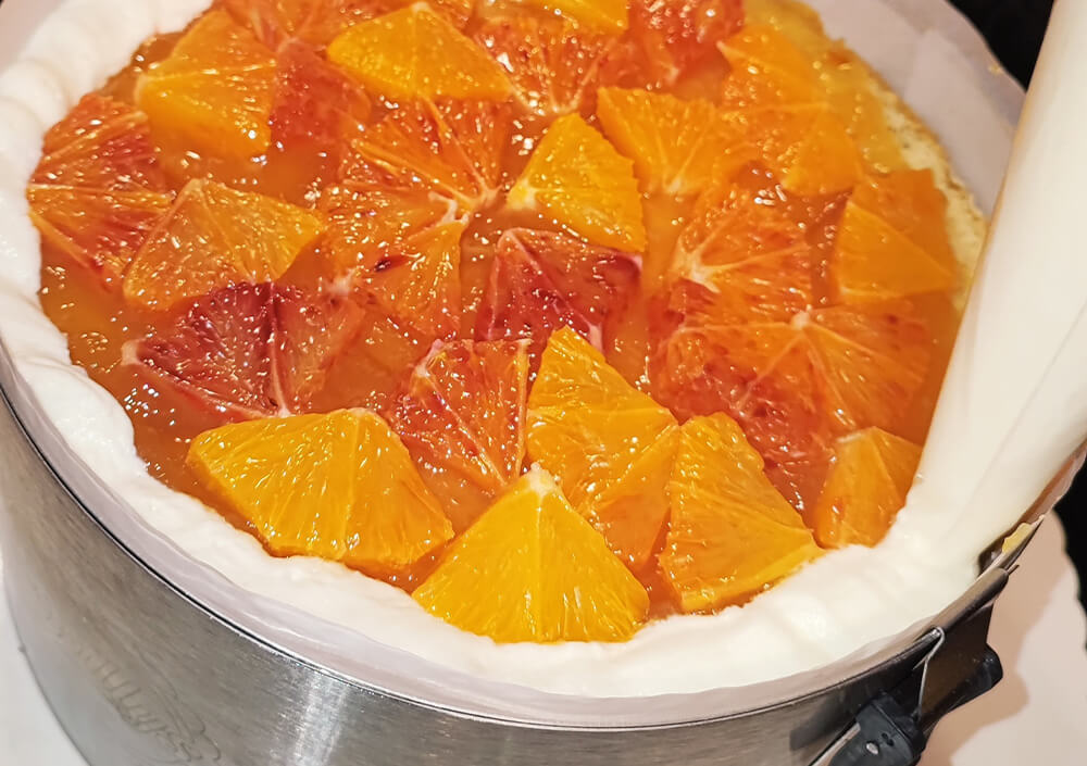 Nahaufnahme eines Käsekuchens in einer Springform, der mit Orangenscheiben und einer glänzenden Glasur überzogen ist. Mit einem Spritzbeutel wird ein weißer Cremerand um den Rand herum aufgetragen.