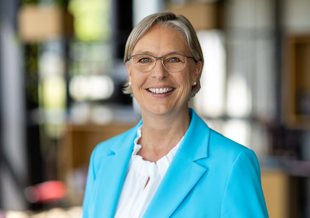 Eine lächelnde ältere Frau mit kurzen grauen Haaren und Brille, die einen hellblauen Blazer und eine weiße Bluse trägt, steht in einem modernen, unscharfen Bürogebäude.