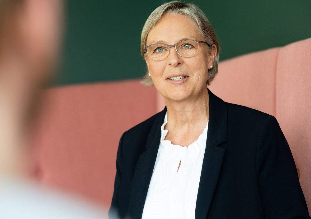 Eine Frau mit kurzem blondem Haar und Brille, die einen schwarzen Blazer und eine weiße Bluse trägt, sitzt und spricht mit einer Person, deren Rücken im Vordergrund zu sehen ist. Sie wirkt engagiert und aufmerksam.