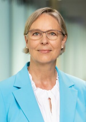Eine Frau mittleren Alters mit kurzen blonden Haaren und Brille, die einen hellblauen Blazer und eine weiße Bluse trägt, posiert vor einem sanft verschwommenen Innenhintergrund.