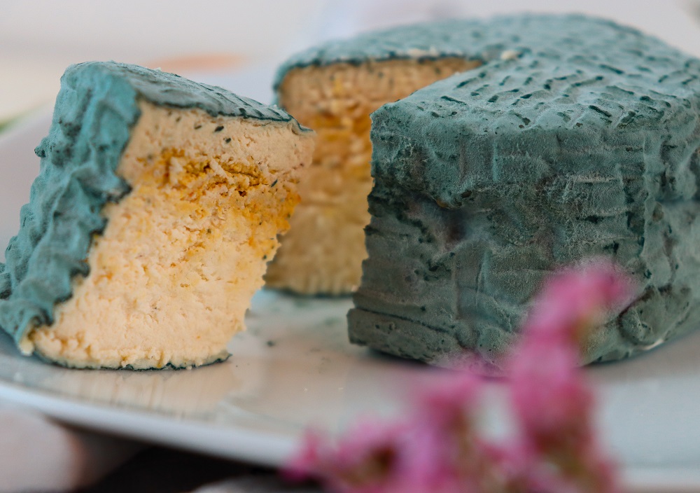 Ein runder Laib der veganen Käsealternative Veeze Oldenburg mit strukturierter Rinde, ein Keil herausgeschnitten und daneben auf einen weißen Teller gelegt. Der Käse hat ein cremiges, hellgelbes Inneres. Im Vordergrund sind rosafarbene Blumen verschwommen zu erkennen.