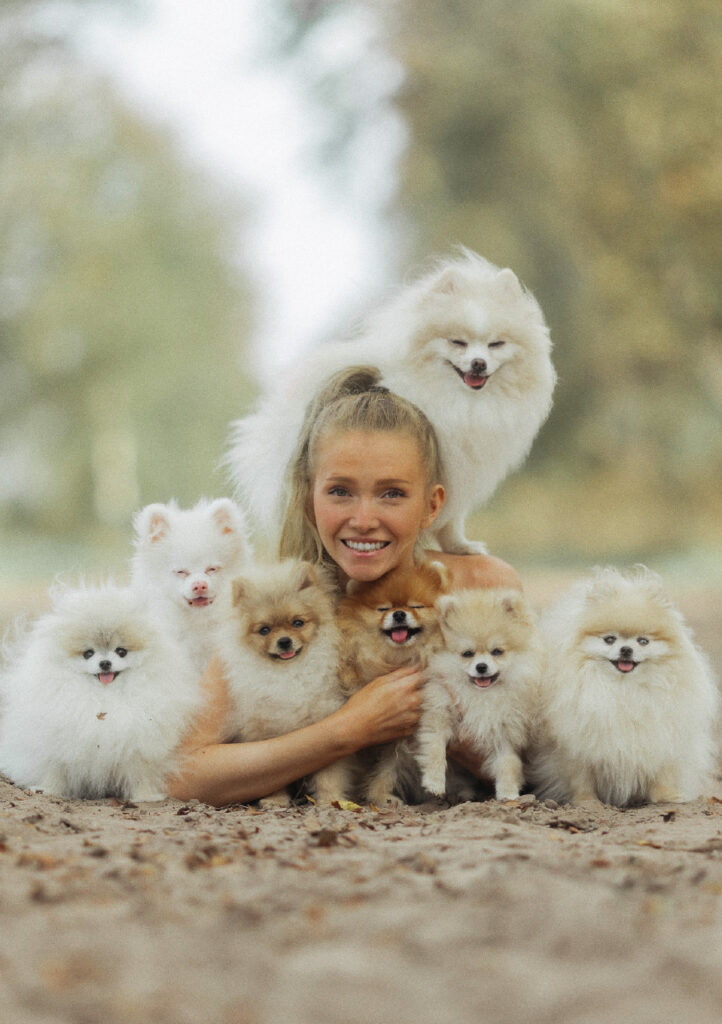 Eine lächelnde Frau sitzt im Freien, umgeben von sechs flauschigen Pomeranian-Hunden, wobei ein Hund auf ihren Schultern sitzt. Der Hintergrund ist unscharf mit Grünzeug.