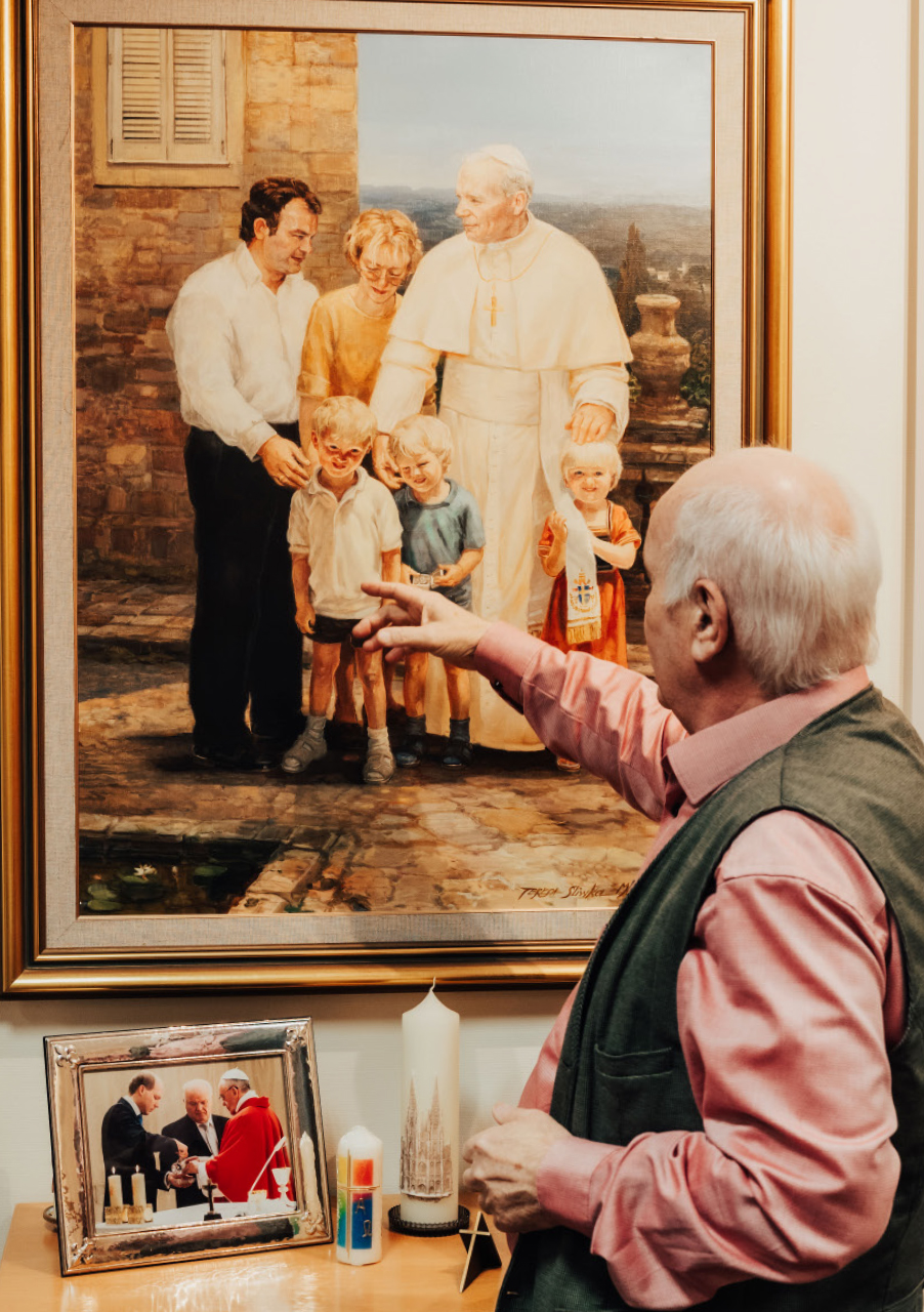 Ein älterer Mann zeigt auf ein gerahmtes Gemälde, das eine Familie mit drei Kindern zeigt, die neben einem Papst steht; unter dem Gemälde befindet sich ein Regal mit einem Foto, einer Kerze und kleinen Gegenständen - auf einem Bild ist angeblich Reiseunternehmer Hans Höffmann zu sehen.