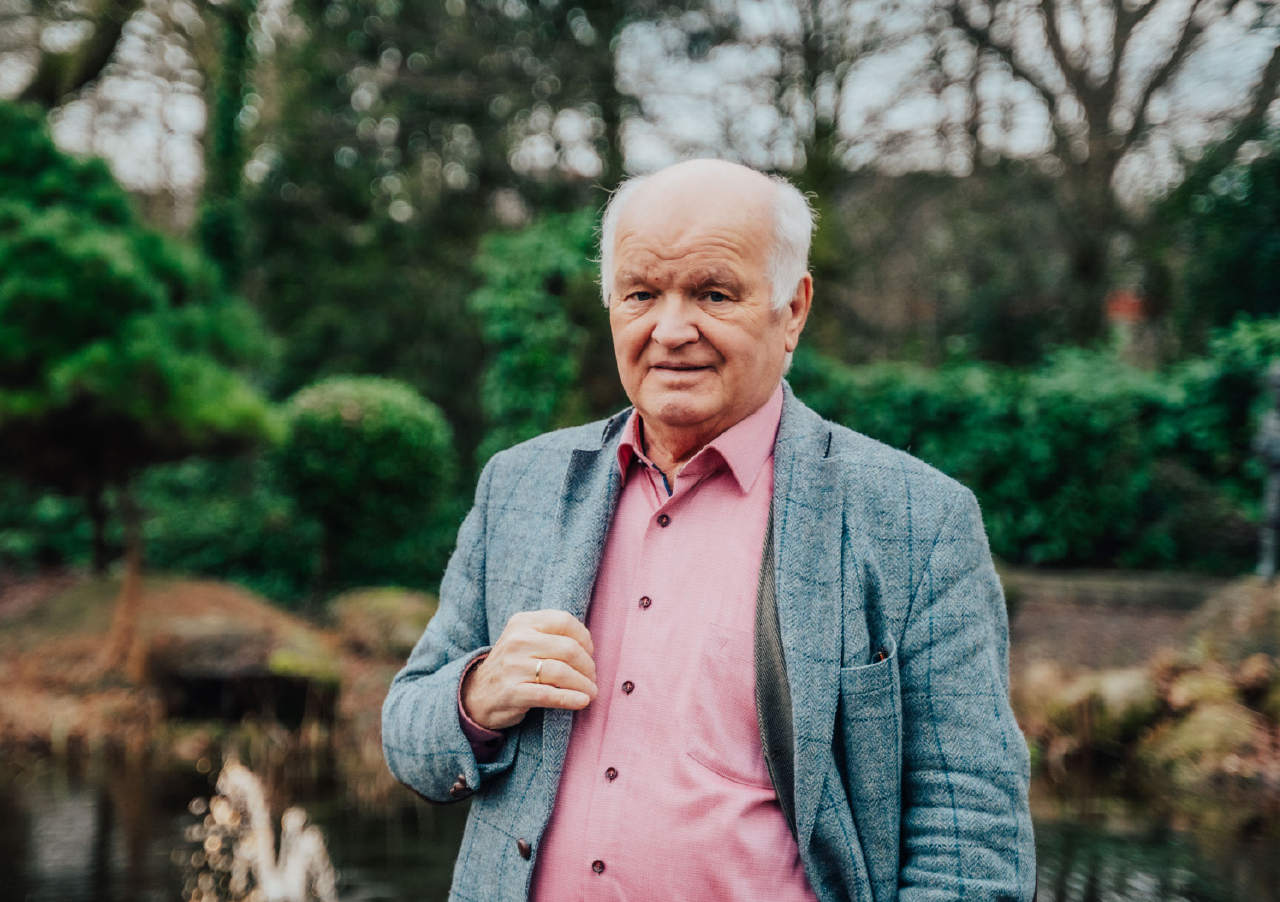 Unternehmer Hans Höffmann, ein älterer Mann mit weißem Haar, steht in einem Park im Freien, trägt ein rosa Hemd und einen grauen Blazer, während im Hintergrund Grünflächen und Bäume verschwimmen.