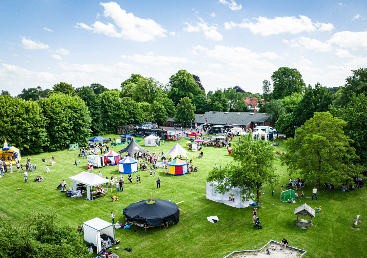 Luftaufnahme eines lebhaften Open-Air-Festivals auf einer Wiese mit bunten Zelten, kleinen Ständen, Menschen, die spazieren gehen und sitzen, und üppigen grünen Bäumen, die das Gebiet unter einem teilweise bewölkten Himmel umgeben.