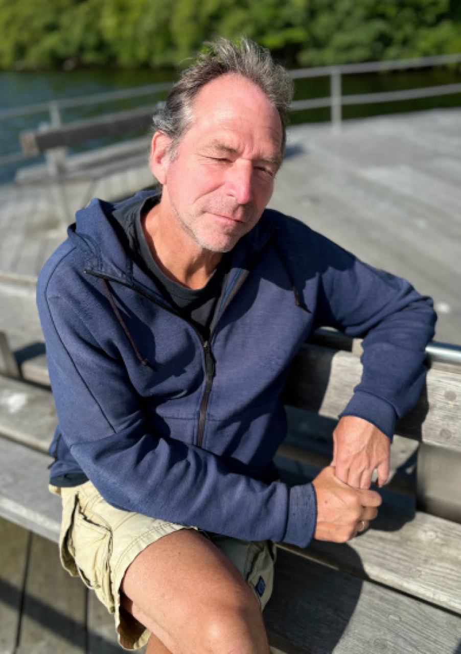 Ein Mann, der einen blauen Kapuzenpullover mit Reißverschluss und beigefarbene Shorts trägt, sitzt im Freien auf einer Holzbank, die Augen geschlossen, und das Sonnenlicht wirft Schatten auf sein Gesicht.