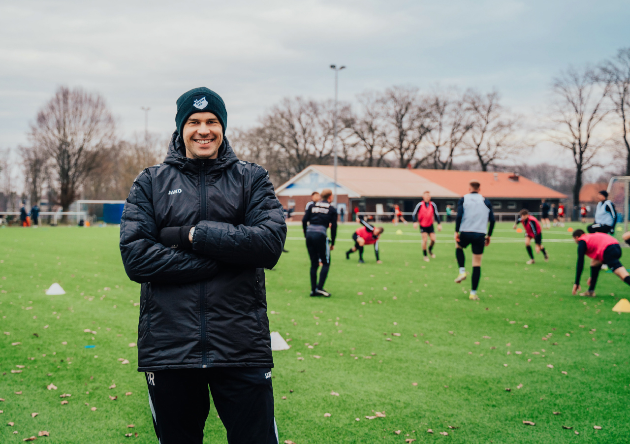 Mitarbeiter Florian Zwick, lächelnd in schwarzer Jacke und Mütze, steht mit verschränkten Armen auf einem grasbewachsenen Fußballplatz, während im Hintergrund mehrere Personen trainieren. Hinter dem Feld sind Bäume und ein Gebäude zu sehen.