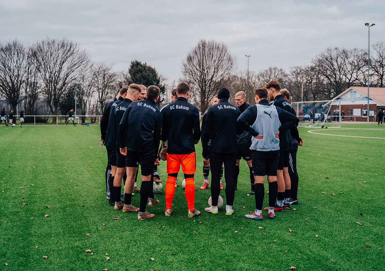 Eine Gruppe von Fußballspielern, trainiert von Florian Zwick: Führungskraft und Fußballtrainer, stehen in dunklen Trikots des SC Bakum auf einem grünen Feld, umgeben von blattlosen Bäumen und einem wolkenverhangenen Himmel.