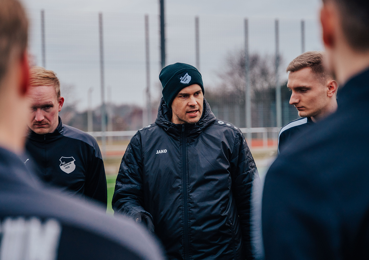 Florian Zwick, Führungskraft und Fußballtrainer, spricht in schwarzer Jacke und Beanie mit seinen Spielern während eines Outdoor-Teammeetings auf dem Feld. Die Spieler hören aufmerksam zu; im Hintergrund sind ein Zaun und Bäume sichtbar.
