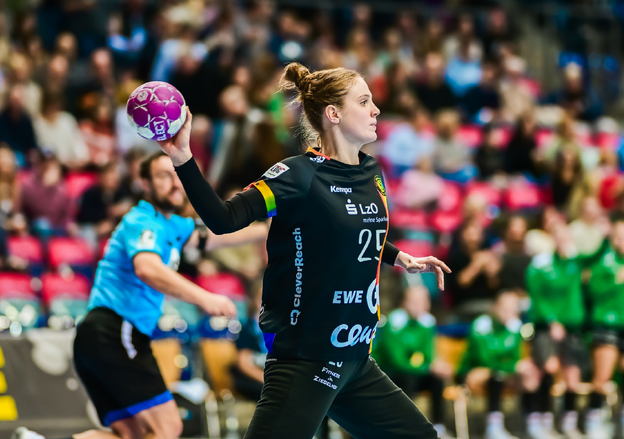 Eine Handballspielerin in einem schwarzen Trikot bereitet sich darauf vor, während eines Spiels einen rosafarbenen Ball zu werfen, während im Hintergrund eine unscharfe Menschenmenge und andere Spieler zu sehen sind.