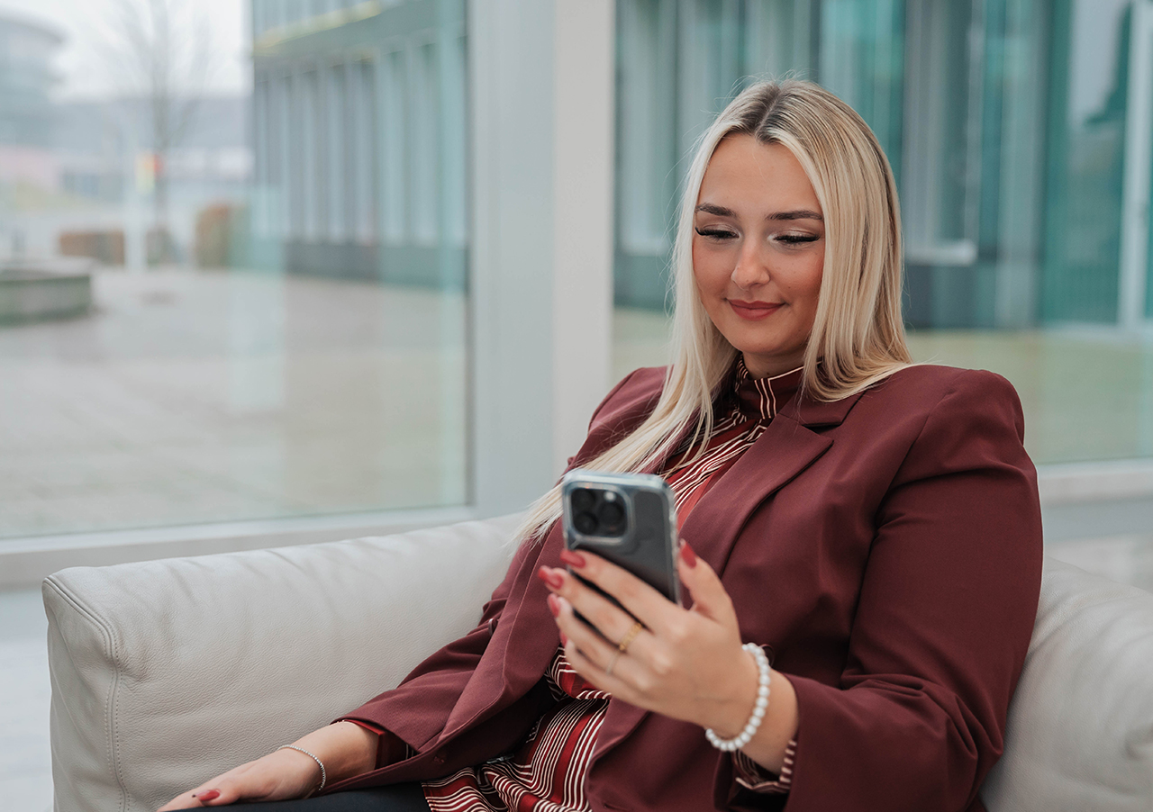 Eine Frau mit langen blonden Haaren und einem kastanienbraunen Blazer sitzt auf einer hellen Couch in einem Haus und lächelt auf ihr Smartphone, während sie digitales Banking erkundet. Im Hintergrund sind große Fenster und moderne Gebäude zu sehen.