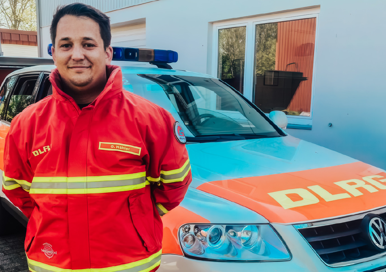 Eine Person in rot-gelber DLRG-Uniform steht an einem sonnigen Tag vor einem Fahrzeug mit DLRG-Kennzeichnung und blauem Notlicht vor einem Gebäude.