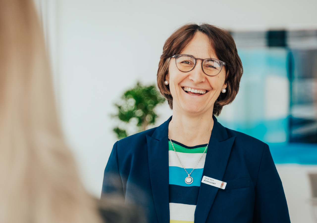 LzO-Vorständin Asmussen, warm lächelnd in blauem Blazer und mit Brille, steht in einem Innenraum für ein Interview. Sie hat kurze braune Haare, trägt ein gestreiftes Hemd und ein Namensschild, während von hinten im Vordergrund eine weitere Person auftaucht.