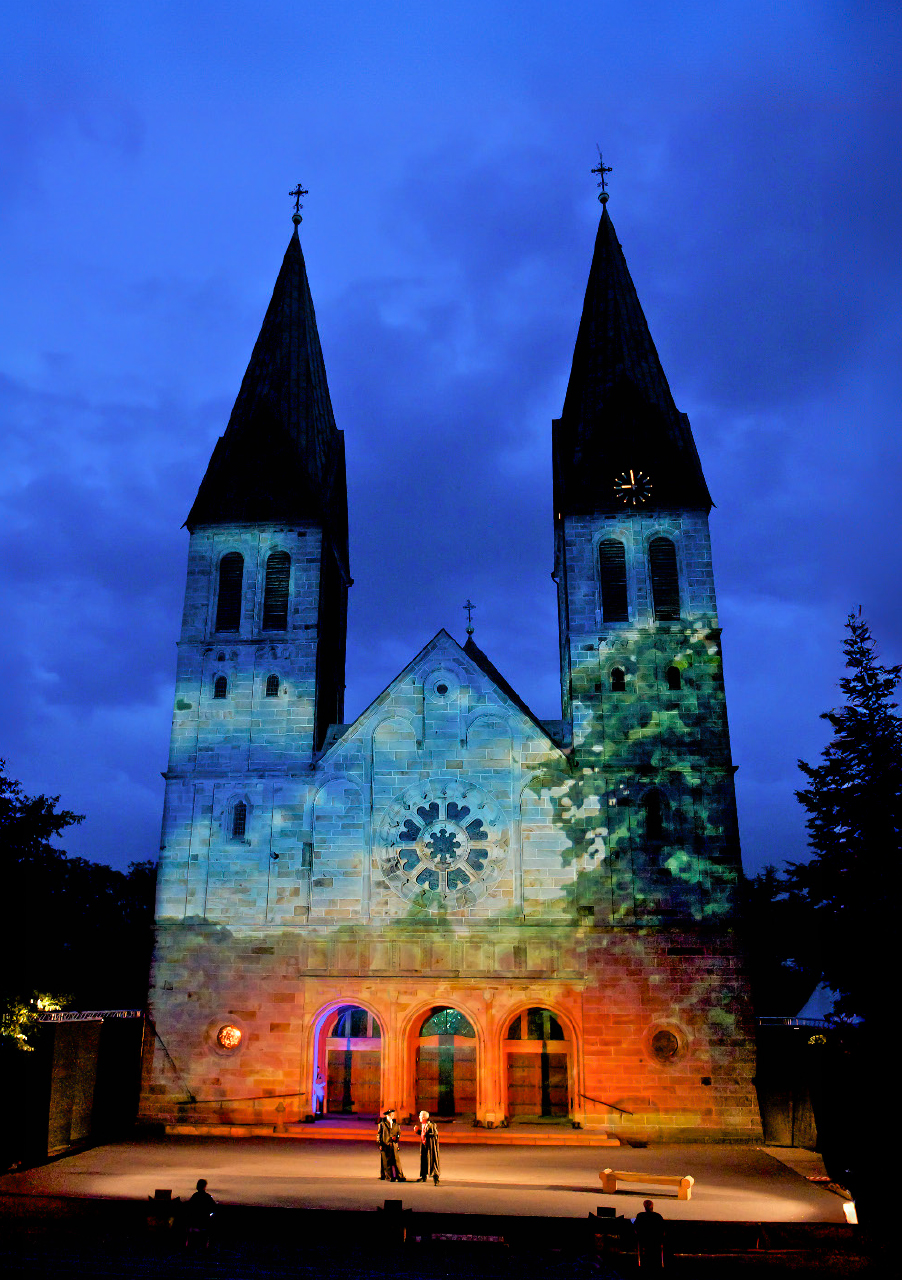 Vor einem tiefblauen Abendhimmel steht eine Kirche mit zwei hohen Türmen, die von bunten Lichtern angestrahlt werden. In der Nähe des Eingangs haben sich einige Menschen versammelt, als warteten sie auf eine Aufführung des Theaters für Jedermann; dramatische Wolken tragen zu der stimmungsvollen Atmosphäre bei.