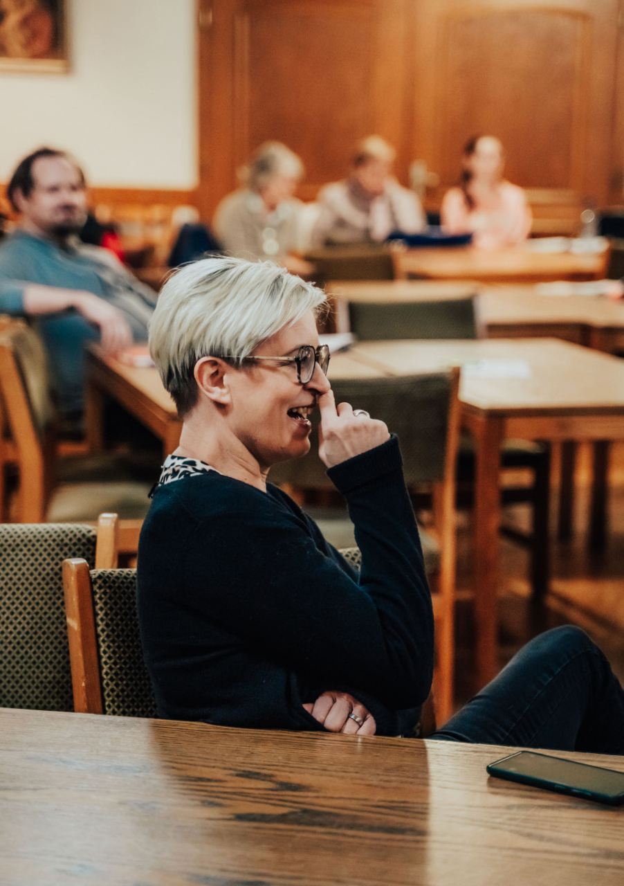Eine Frau mit kurzem blondem Haar und Brille sitzt an einem Holztisch und hält sich lächelnd die Hand vors Gesicht. Im Hintergrund versammeln sich mehrere Personen in einem Raum mit holzgetäfelten Wänden, was auf ein intimes Theater für Jedermann schließen lässt.