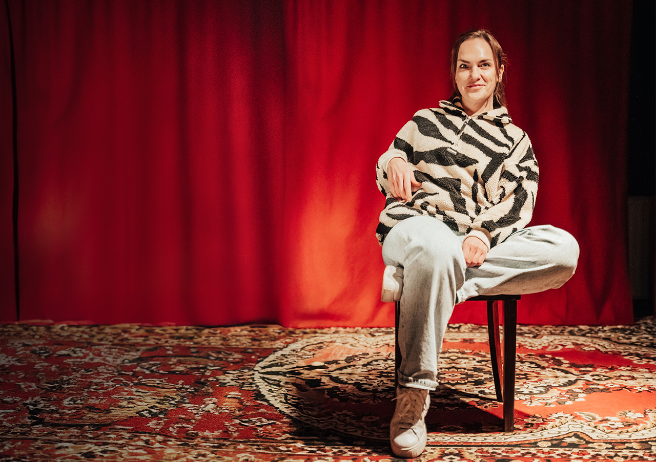 Die Comedian Ulli Lissner, die einen zebragestreiften Pullover und eine helle Jeans trägt, sitzt im Schneidersitz auf einem Stuhl über einem gemusterten Teppich vor einem roten Vorhang, lächelt leicht und wirkt entspannt.