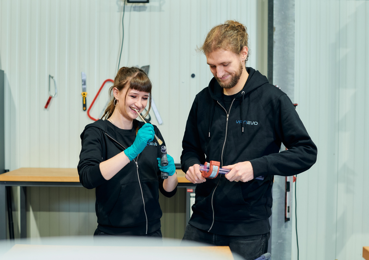 Zwei Personen in schwarzen Kapuzenpullovern arbeiten zusammen in einer Werkstatt. Einer hält ein Werkzeug und lächelt, der andere hält ebenfalls ein Gerät und blickt nach unten, beide scheinen mit einer gemeinsamen Aufgabe beschäftigt zu sein.