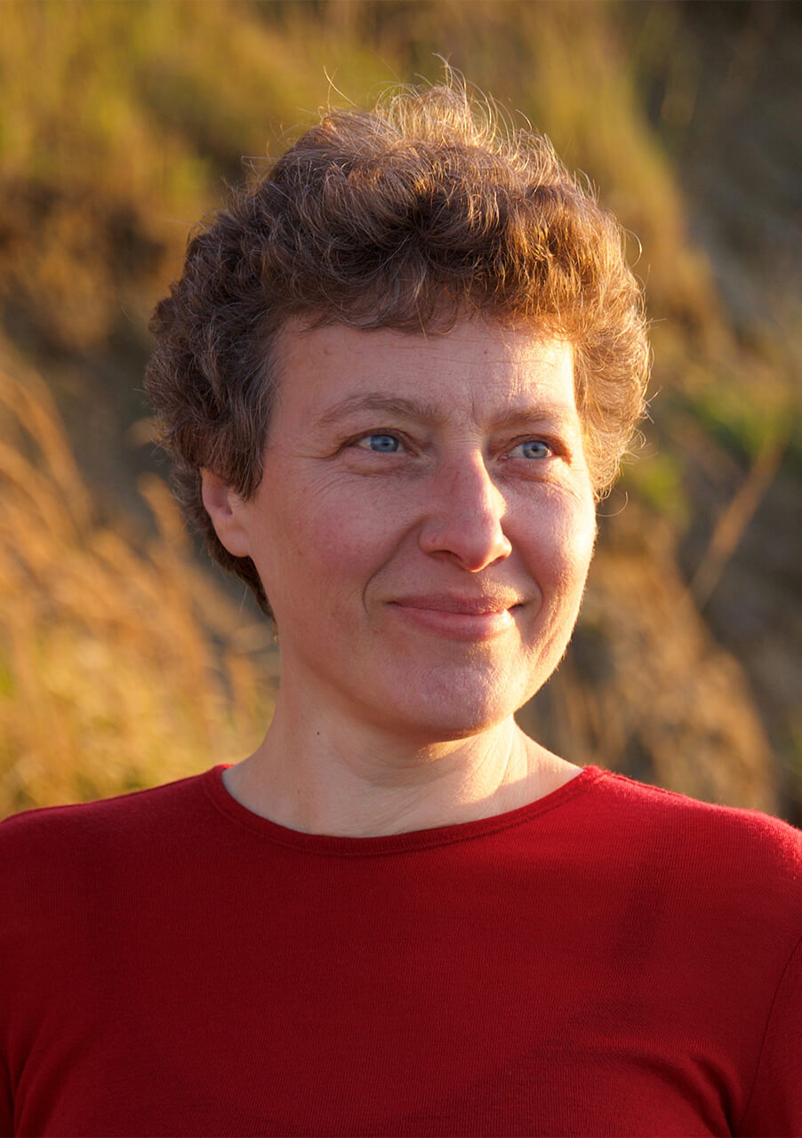 Eine Person mit kurzen, lockigen braunen Haaren und blauen Augen lächelt sanft, während sie im Freien steht und ein rotes Hemd trägt. Der Hintergrund ist sanft mit grünen und goldenen Tönen verschwommen, was an Sonnenlicht auf Gras oder Laub erinnert.