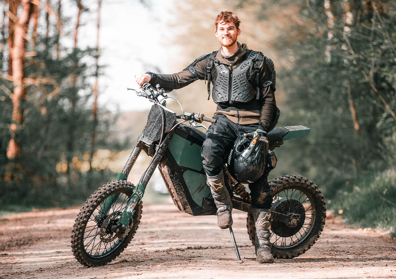 Ein Mann in Schutzkleidung sitzt auf einem Dirt Bike auf einem Waldweg, hält einen Helm und lächelt in die Kamera. Im Hintergrund sind Bäume und Grünflächen zu sehen.