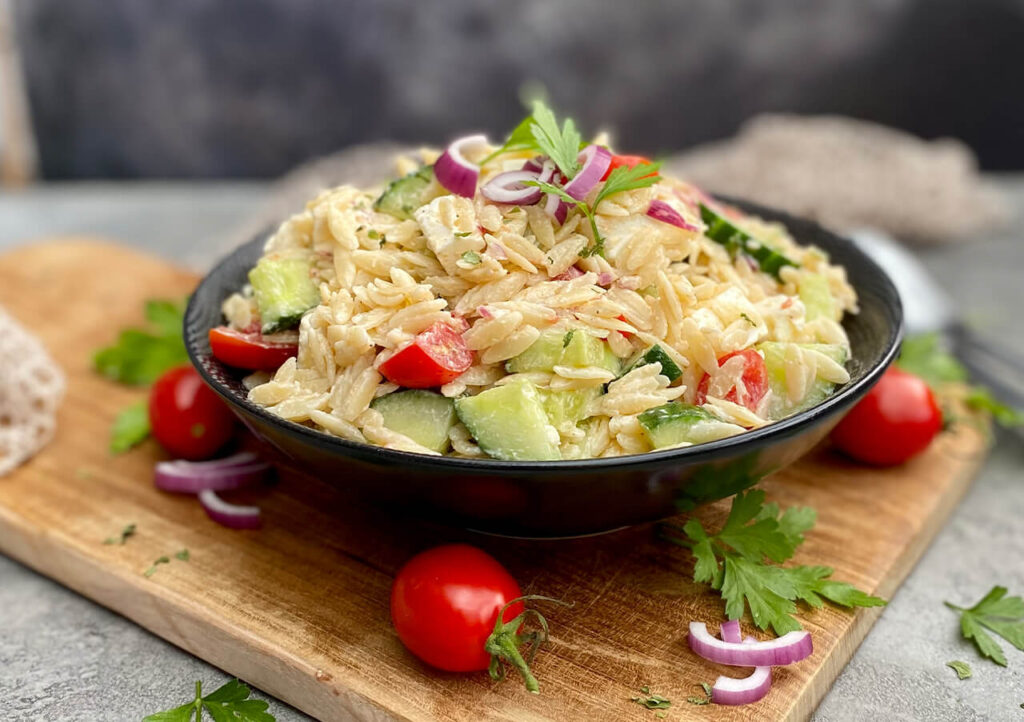 Eine schwarze Schale mit Orzo-Nudelsalat mit geschnittenen Gurken, Kirschtomaten, roten Zwiebeln und Petersilie steht auf einem Holzbrett, auf dem frische Kräuter und Kirschtomaten verstreut sind.