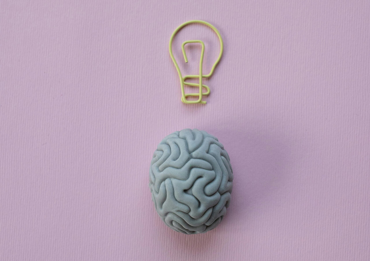 Das Modell eines Gehirns steht auf einem rosafarbenen Hintergrund, darüber befindet sich eine gelbe Büroklammer in Form einer Glühbirne und symbolisiert "Gehirn verstehen und Denken lenken" für mehr Kreativität und Ideen.